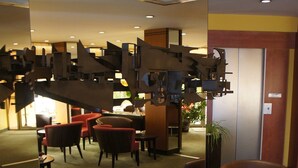 Un vestíbulo de hotel con sillones tapizados en rojo y marrón, mesitas pequeñas y una gran instalación de arte abstracto en el techo.