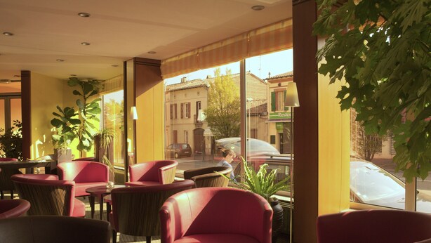 Un café con sillas y mesas rojas, amplios ventanales con vistas a la calle y plantas de interior.