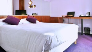 Habitación de hotel con una cama grande, un escritorio, una silla, un cubo de basura y una lámpara fijada a la pared.