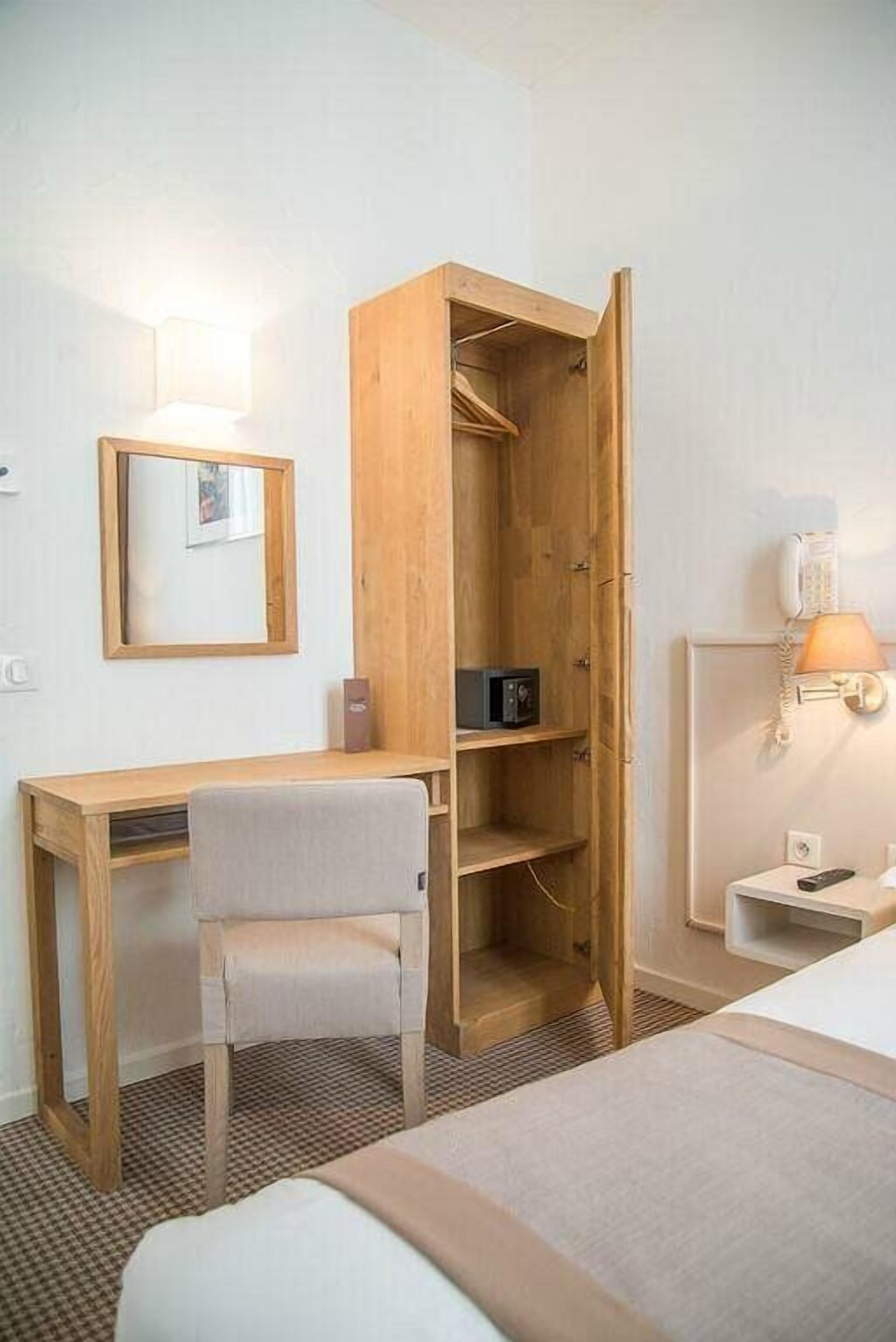 Een hotelkamer met een houten bureau, een stoel, een bed, een kledingkast, een spiegel, een lamp en een telefoon.