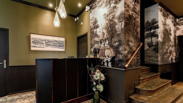 Een receptie met een houten toonbank, een ingelijst schilderij aan de muur, een trap en een muurschildering van een tropisch landschap.
