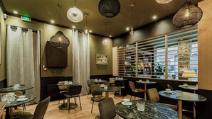 Een modern restaurant met ronde tafels en stoelen, hangende verlichting en uitzicht door een raam met jaloezieën.