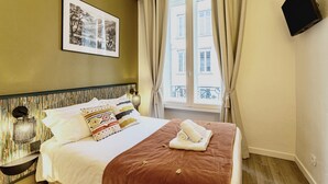 Een hotelkamer met een bed, een inlijsting foto aan de muur, een aan de muur bevestigde televisie en een raam met gordijnen.