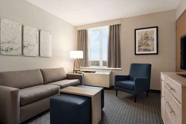 A hotel room with a sofa, a small table, a chair, a desk, a lamp, a TV, and artwork on the walls. - Homewood Suites by Hilton University City (Philadelphia)