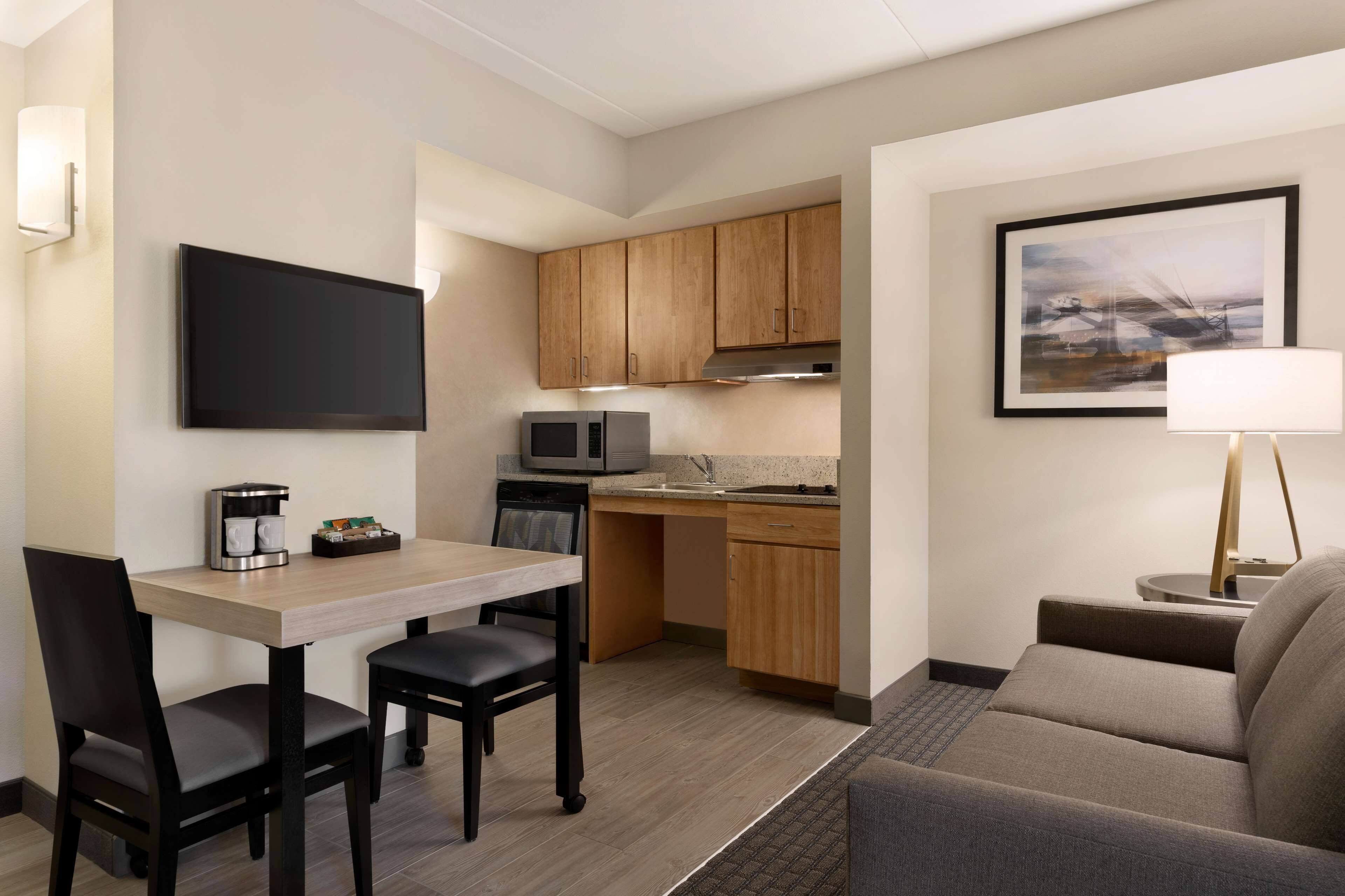 Une chambre d’hôtel compacte comprenant un coin cuisine, un espace repas avec une table et des chaises, un canapé, un téléviseur à écran plat fixé au mur et un tableau encadré.