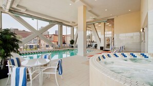 Una piscina interior con sillones de descanso y un jacuzzi, rodeada de amplias ventanas y vistas a edificios.