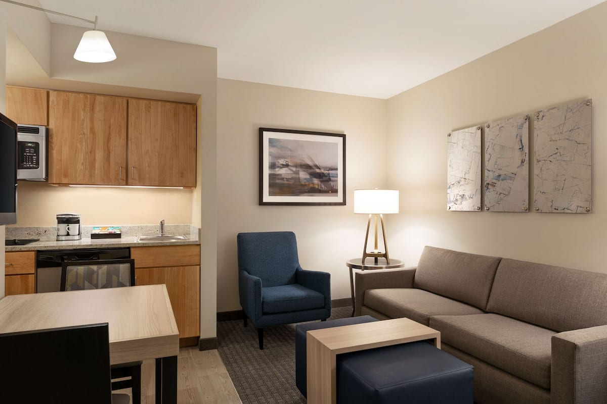 Une chambre d’hôtel compacte comprenant un coin cuisine, un fauteuil bleu, un canapé, une petite table et une lampe.