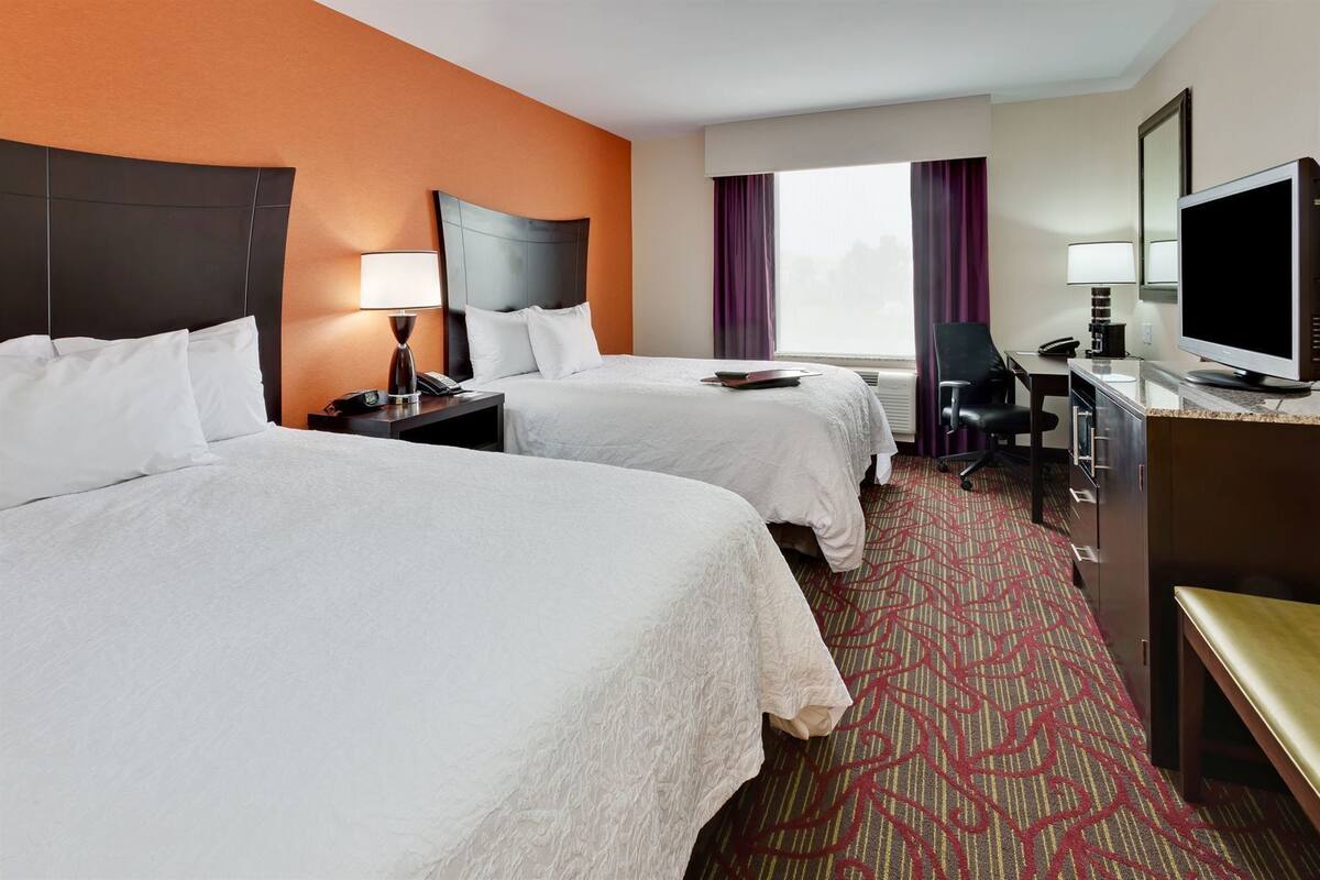 A hotel room with two beds, a desk, a TV, and a window with curtains.