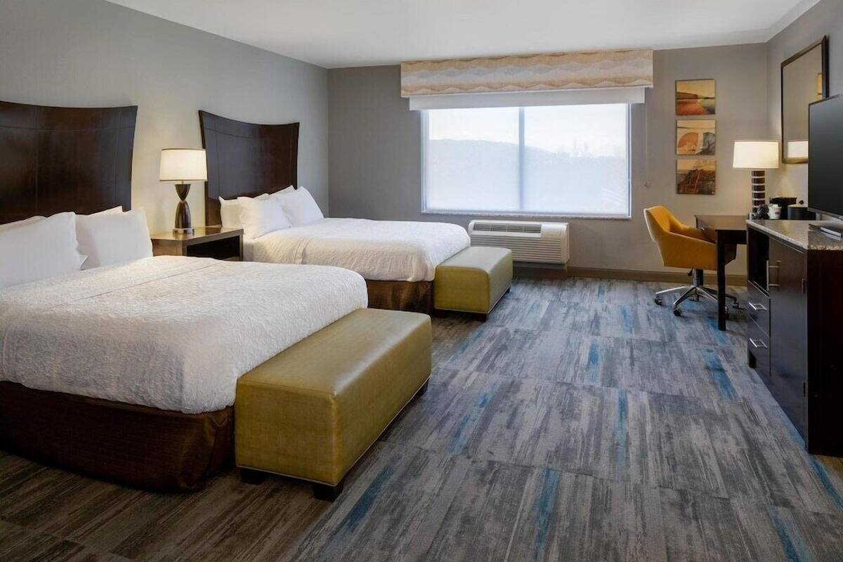 A hotel room with two beds, a desk, a chair, a TV, and a large window.