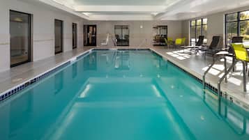 Uma grande piscina interior com água cristalina, rodeada por espreguiçadeiras e uma área iluminada pelo sol com grandes janelas.