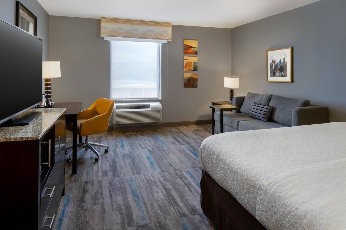 A hotel room with a bed, a desk with a computer, a chair, a sofa, a TV, a window with a view, and a wall with artwork.