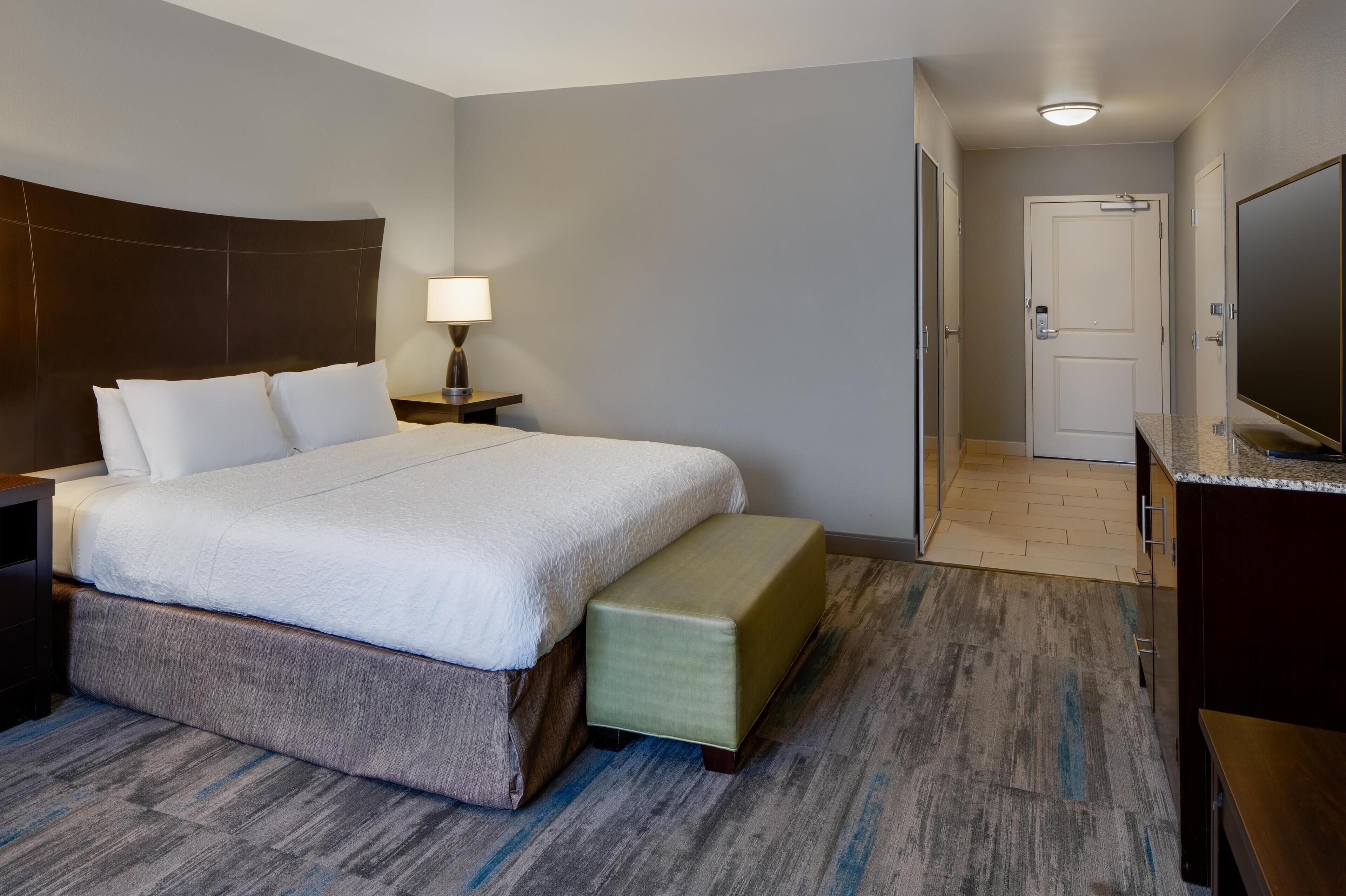 A hotel room with a large bed, a bedside table with a lamp, a bench, a TV, and a door leading to another room.