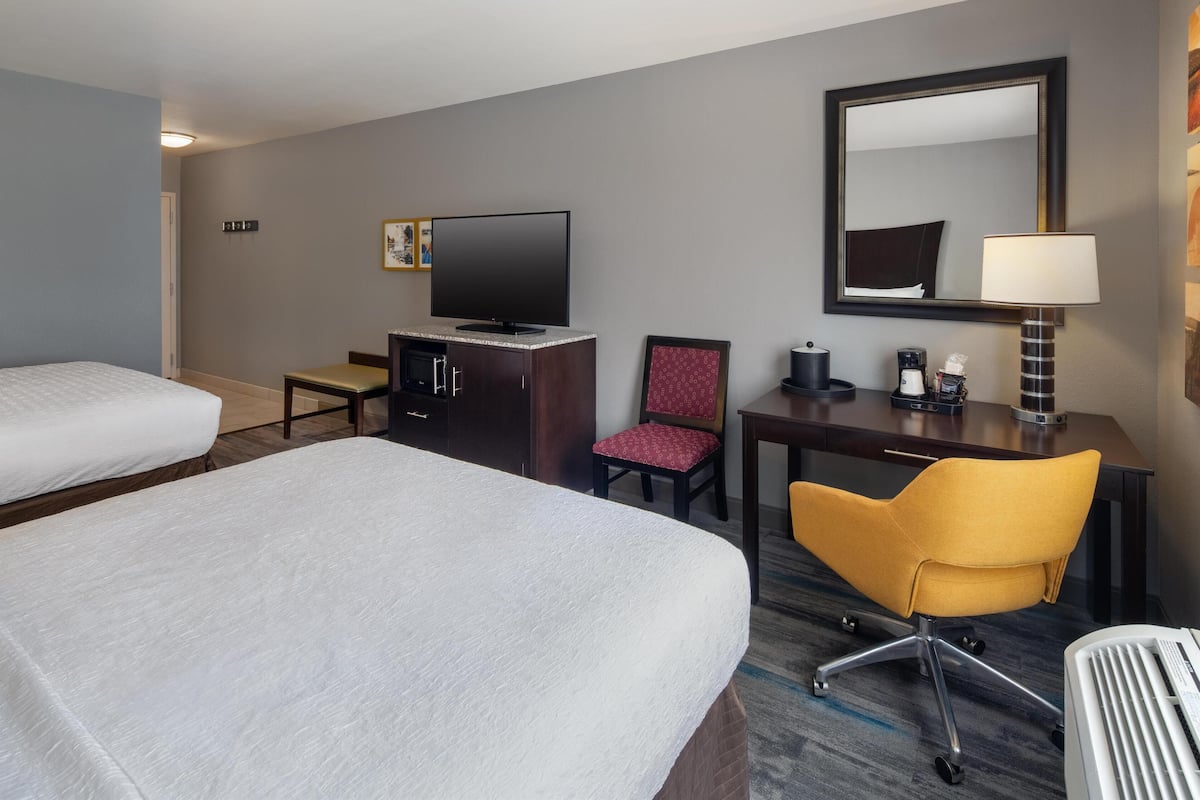 A hotel room with two beds, a desk with a chair, a television, a desk lamp, and a mirror.