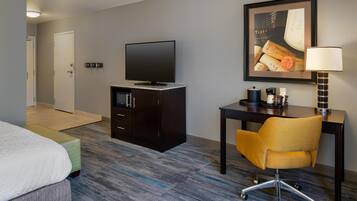 A hotel room with a bed, a desk with a chair, a television, a framed picture on the wall, and a lamp.