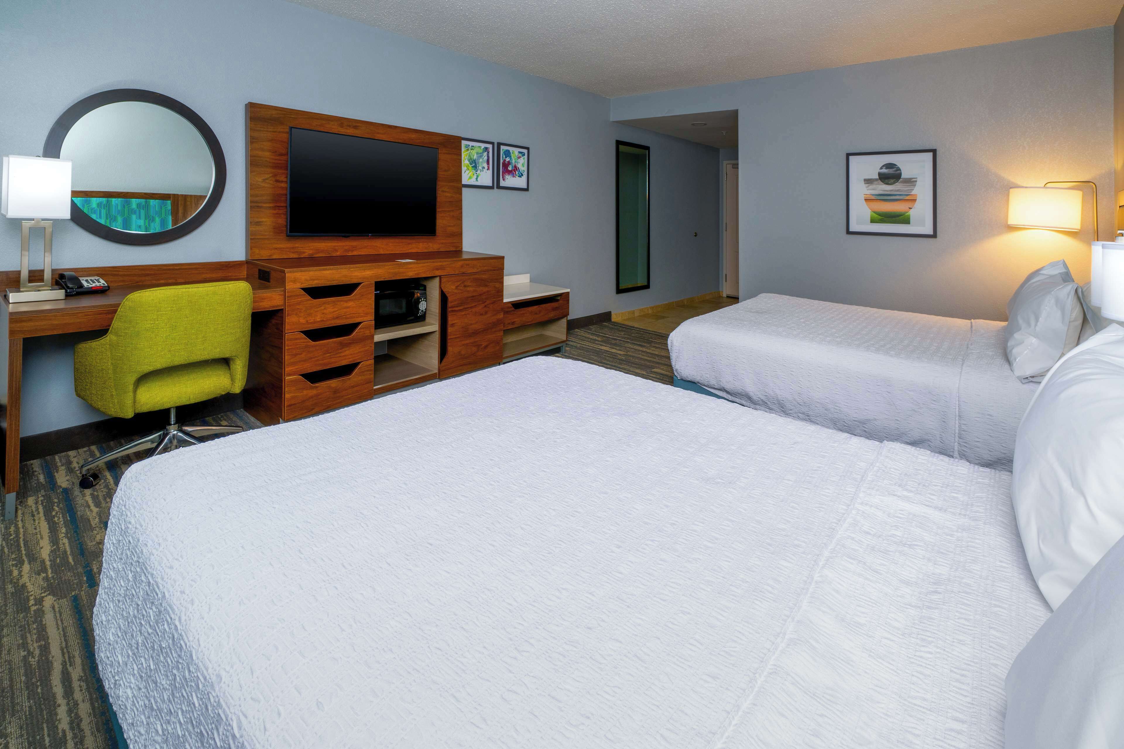 A hotel room with two beds, a desk with a chair, a flat-screen TV, and artwork on the walls.