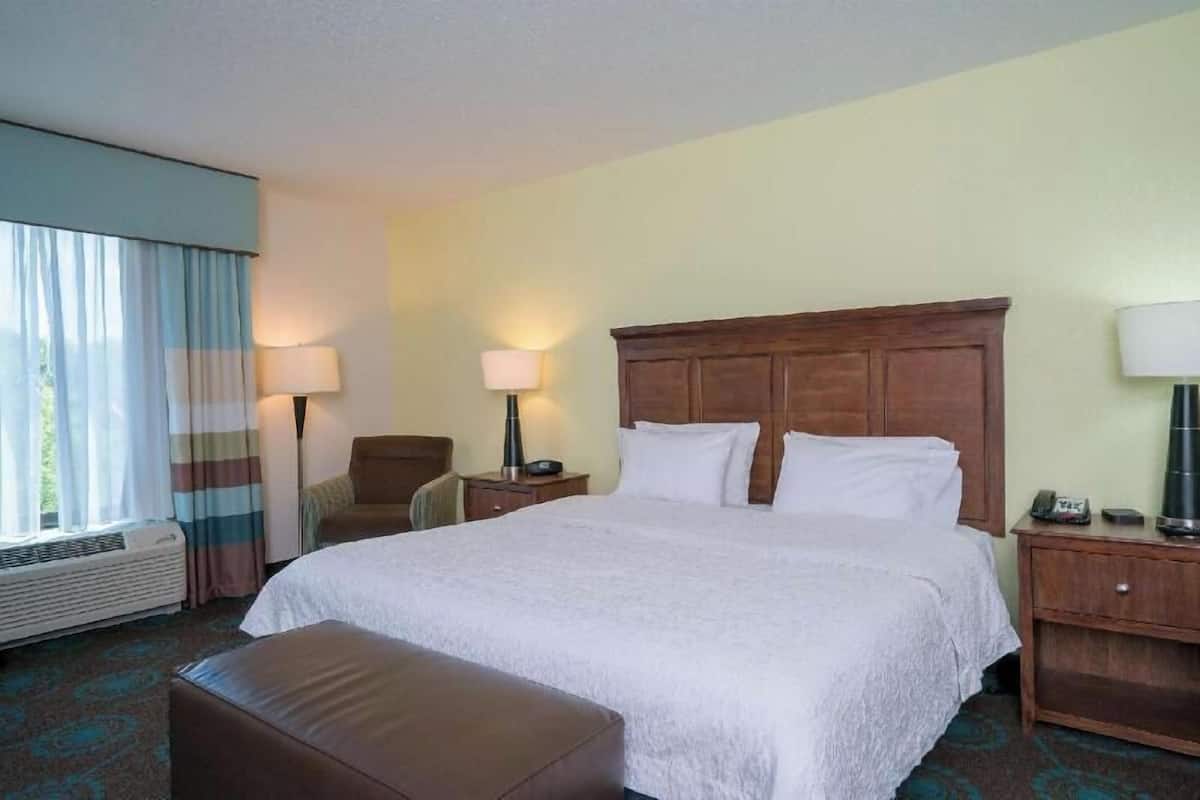 A hotel room with a large bed, two bedside tables, a chair, a window with curtains, and a wall-mounted lamp.