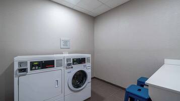 Un cuarto de lavandería con dos lavadoras apiladas, un letrero que indica que hay productos de lavado disponibles para comprar, y un taburete azul pequeño.