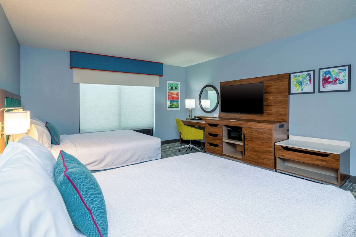 A hotel room with two beds, a desk, a TV, and a window.