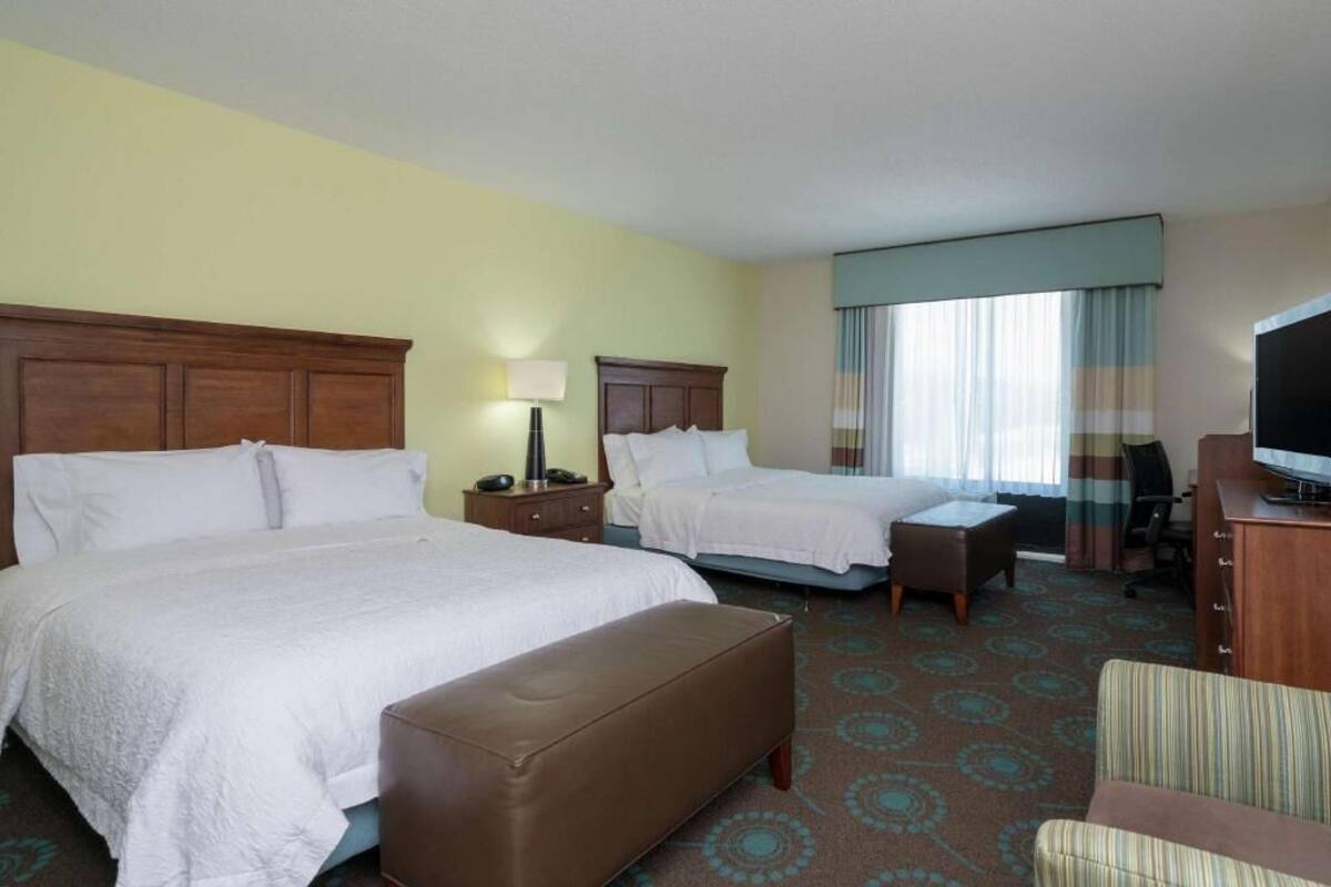 A hotel room with two beds, a bench, a desk with a TV, and a window with curtains.