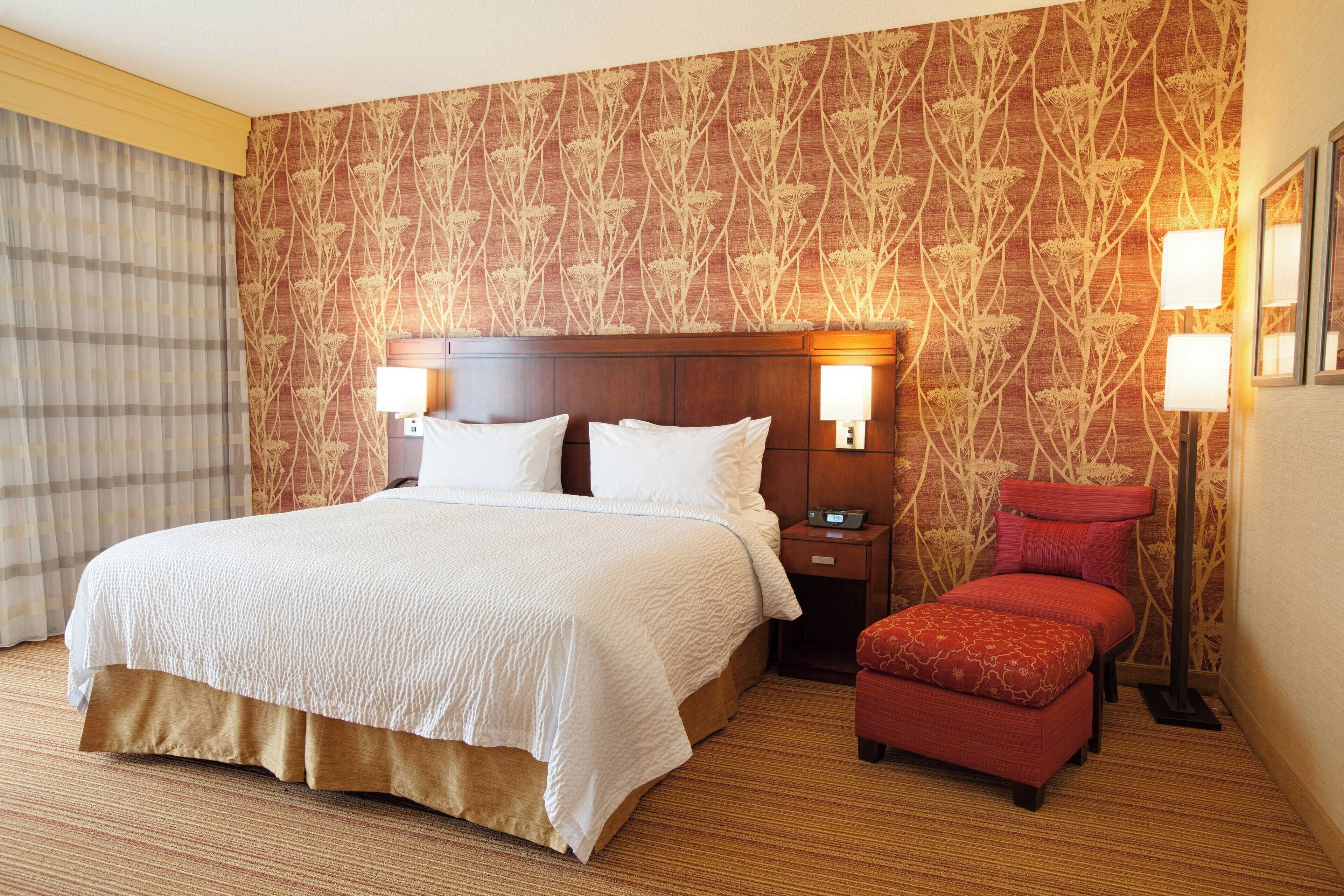 A hotel room with a large bed, a bedside table, a red ottoman, and a patterned wallpaper.
