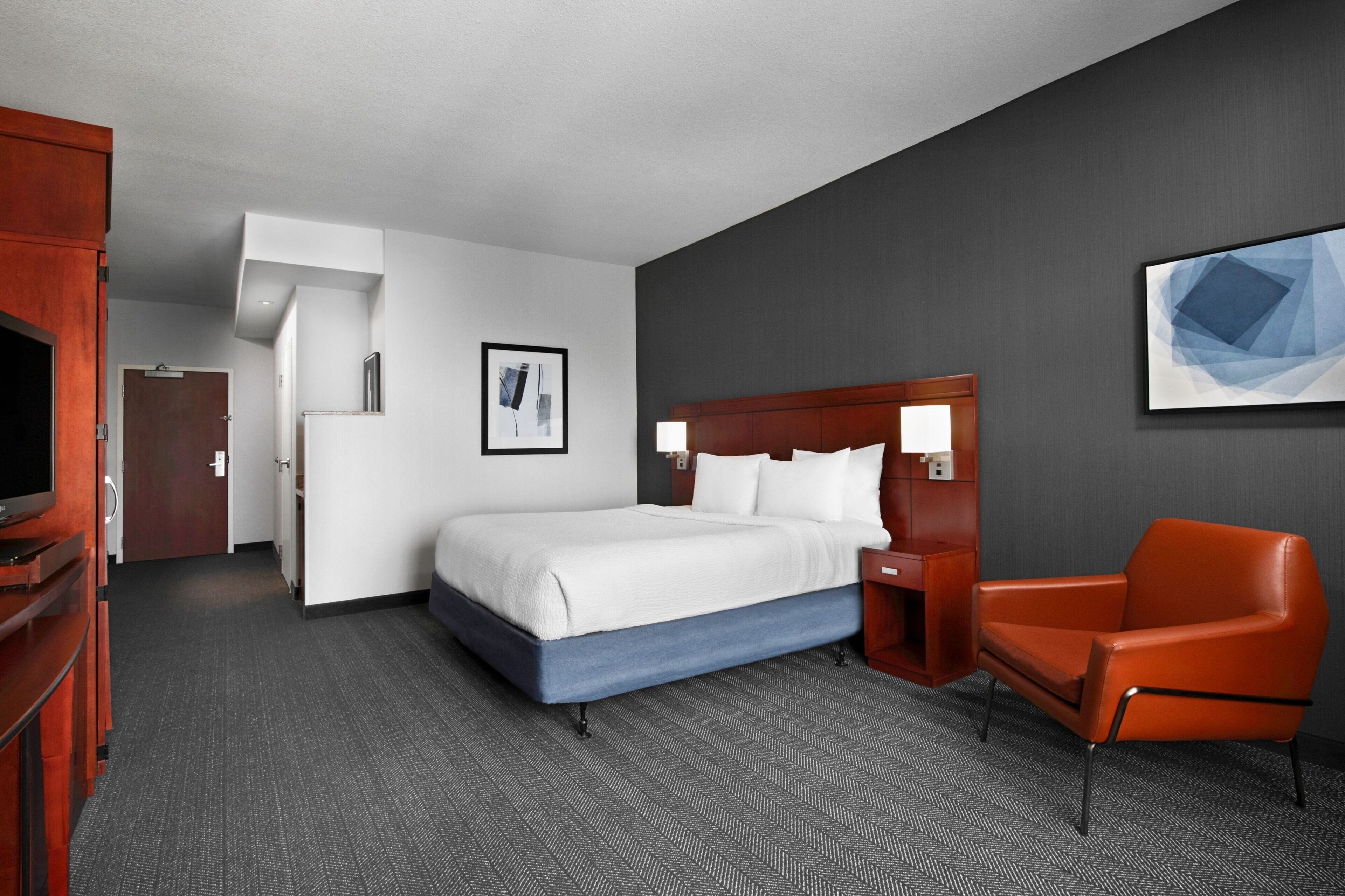 A hotel room with a bed, a chair, a television, and a painting on the wall.