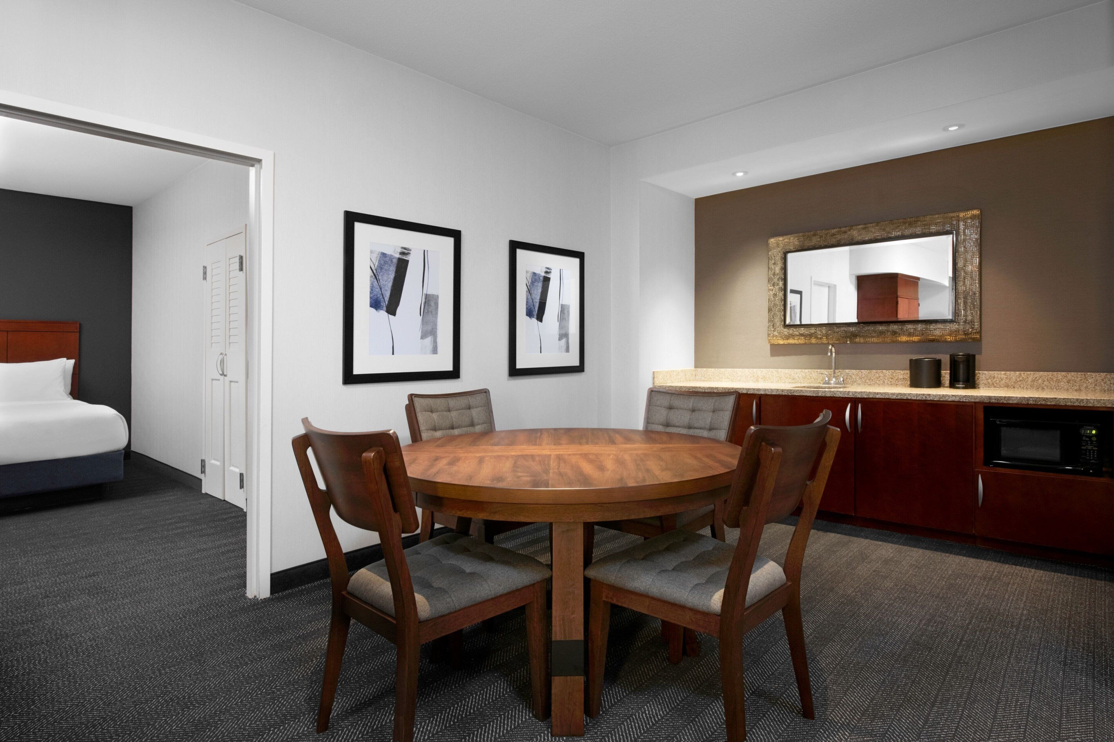 Habitación de hotel con una mesa redonda de madera, cuatro sillas, una cama, una pequeña cocina y dos cuadros en la pared.