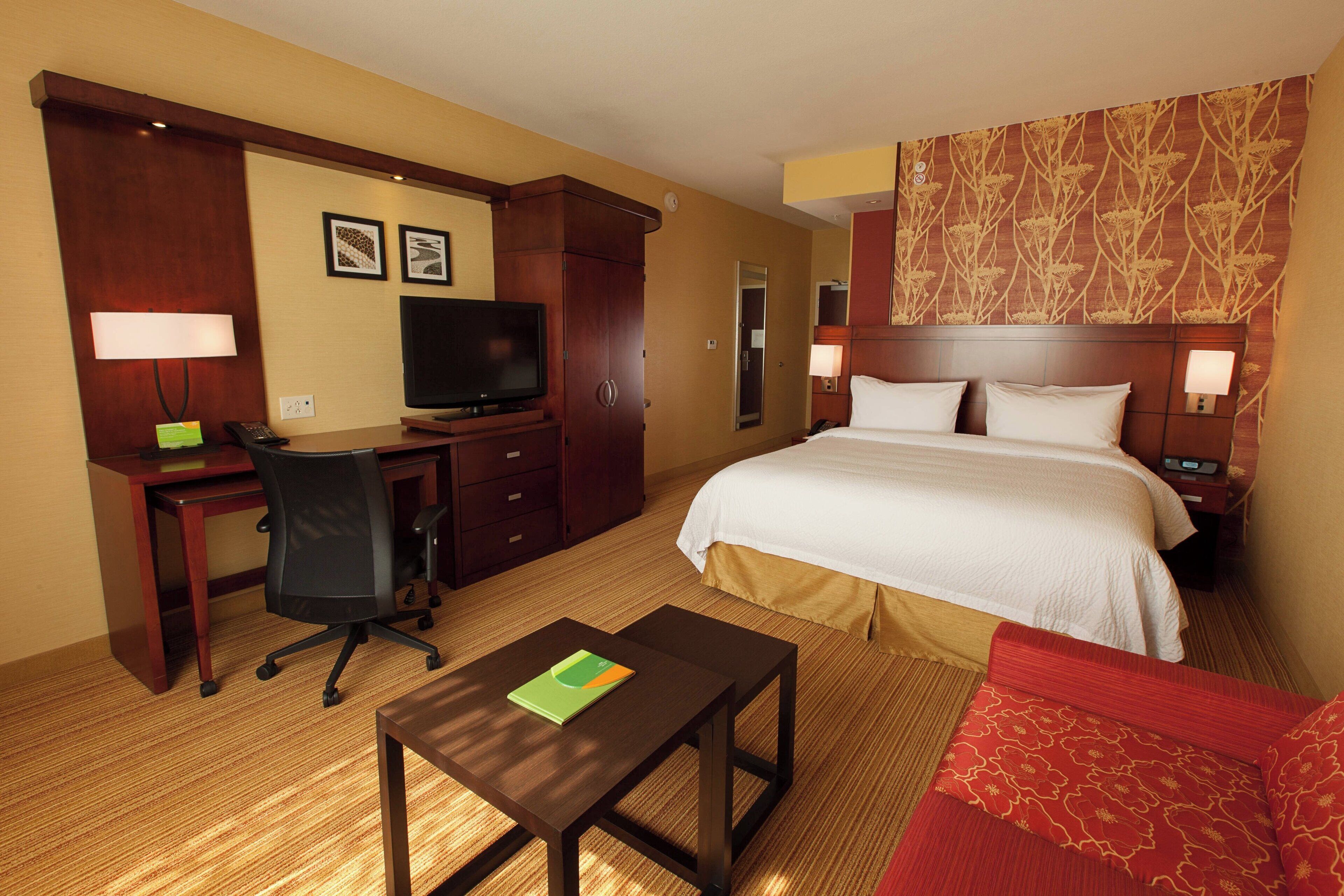 Quarto de hotel com cama, escrivaninha, cadeira, televisão e sofá vermelho.