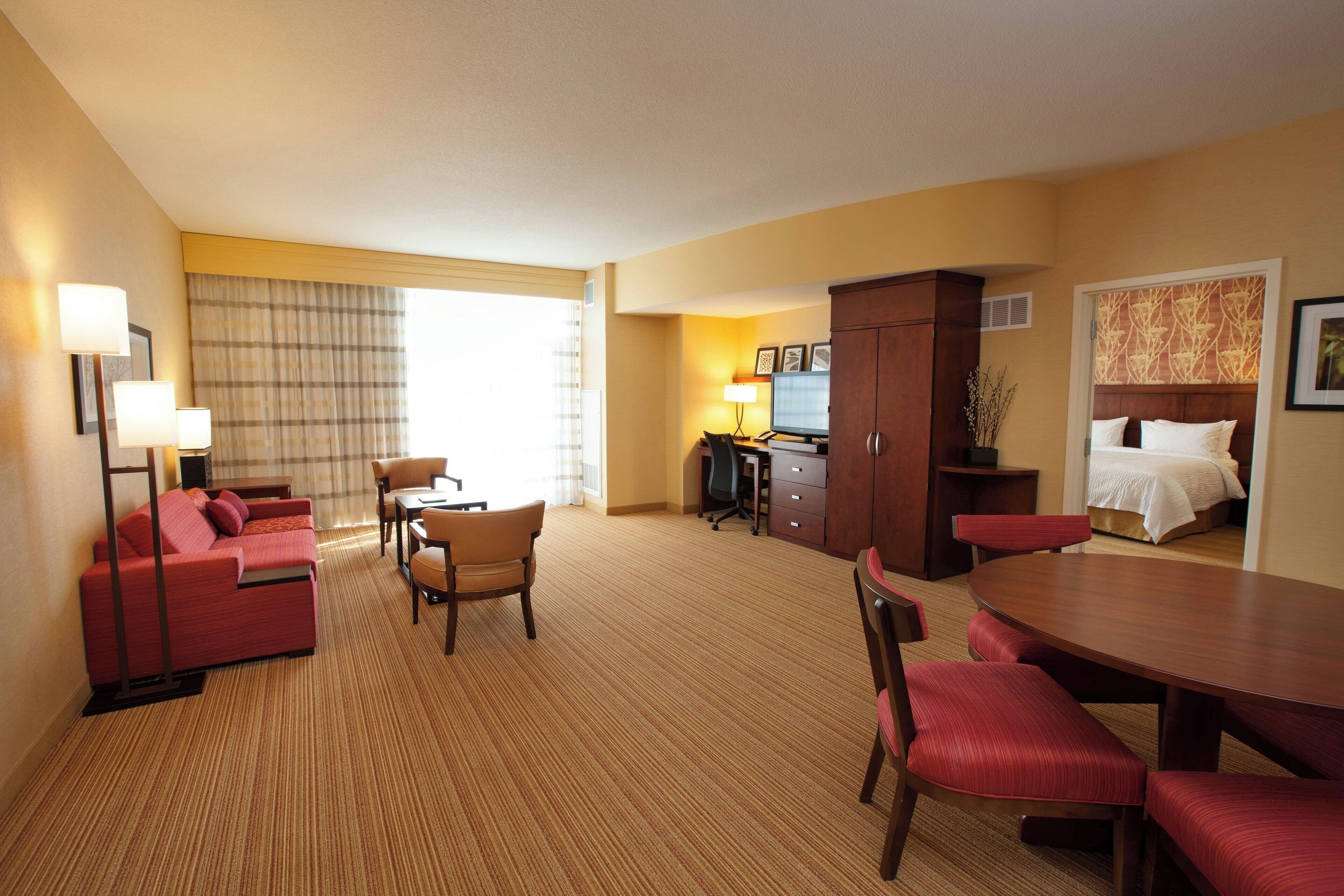 Habitación de hotel con un sofá rojo, una mesa de comedor con sillas rojas, un escritorio con computadora, una cama con ropa blanca y un ventanal con cortinas.