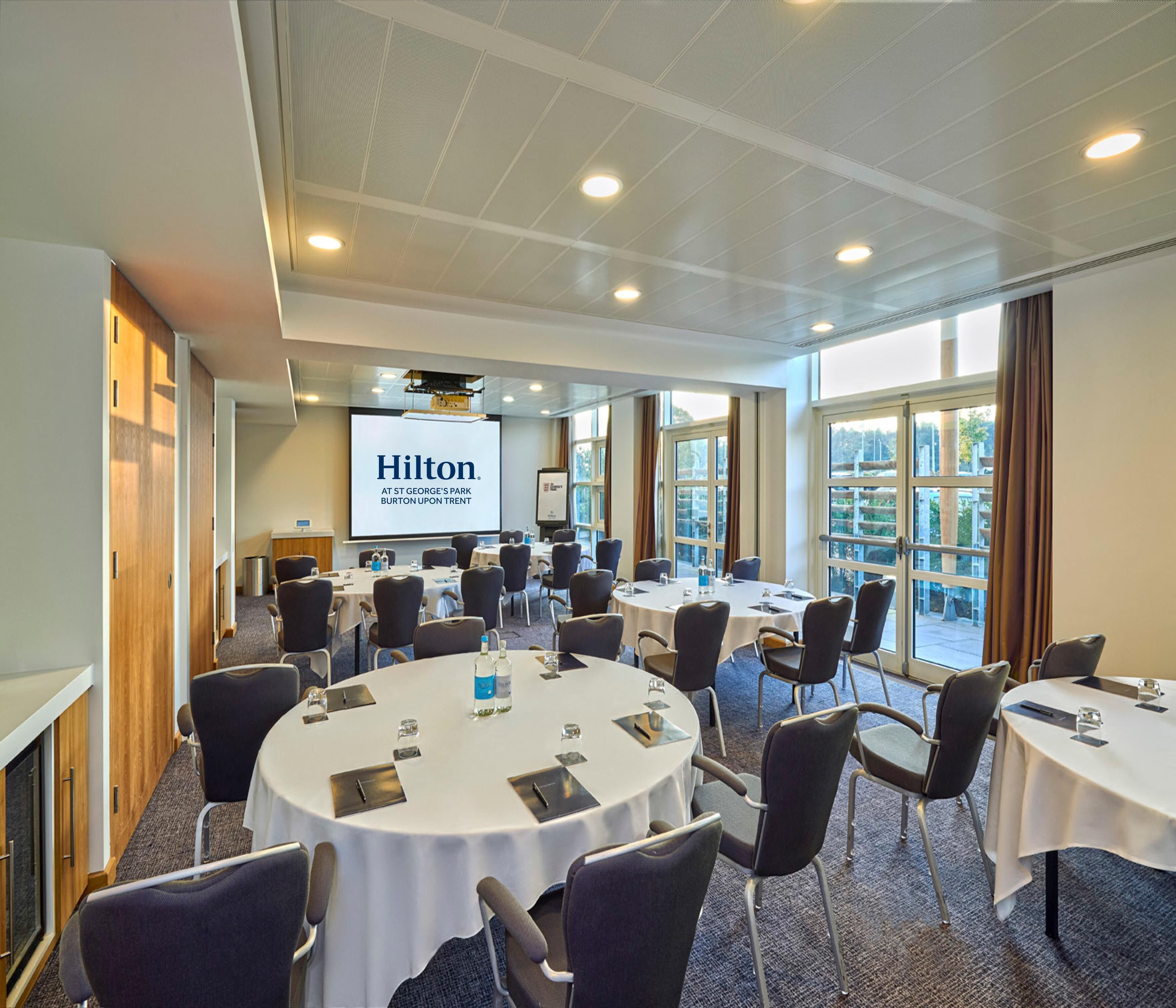 A conference room with multiple tables and chairs, a projector screen displaying 'Hilton at St. George's Park Burton Upon Trent,' and large windows offering a view.