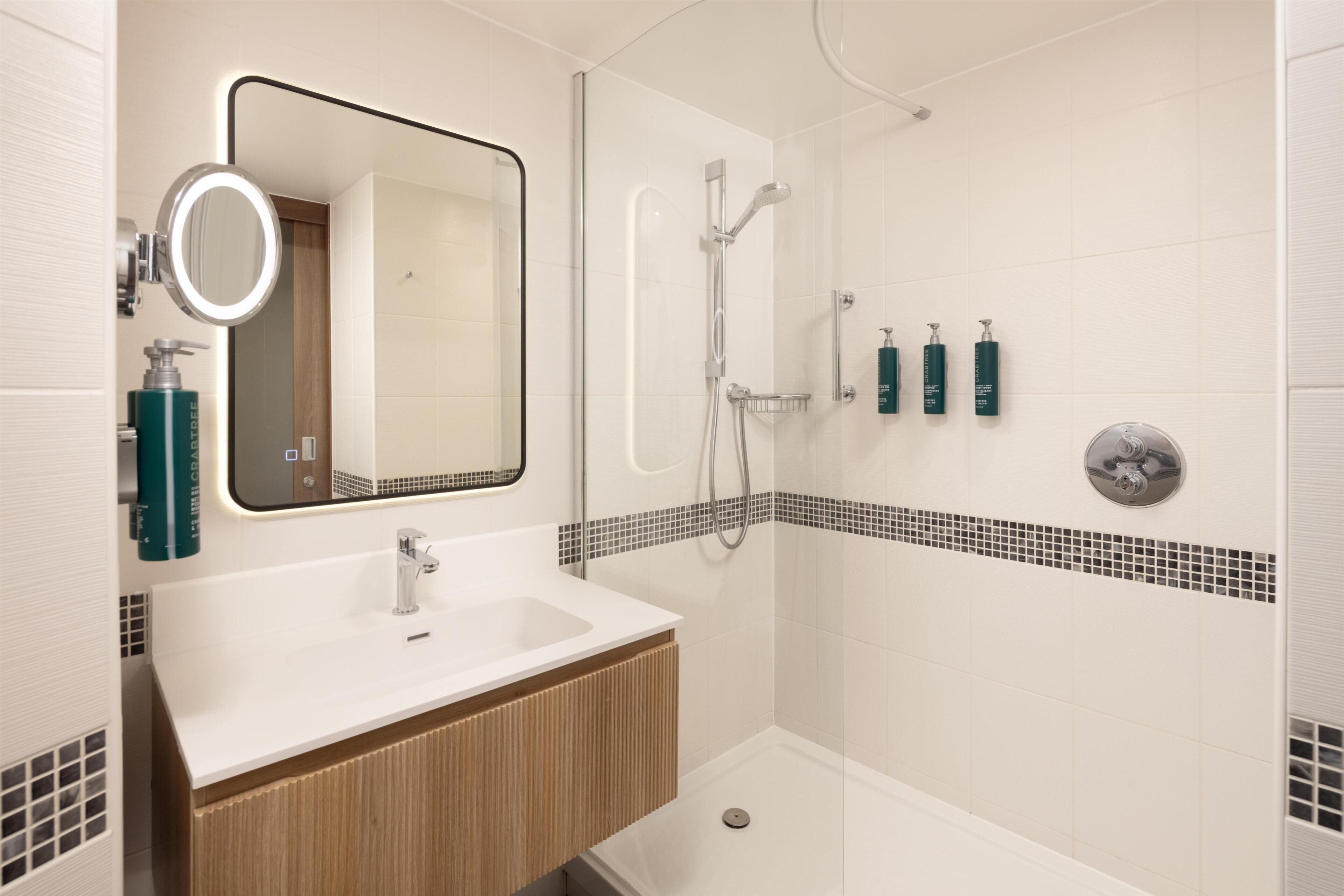 A bathroom with a sink, mirror, shower, and toiletries.