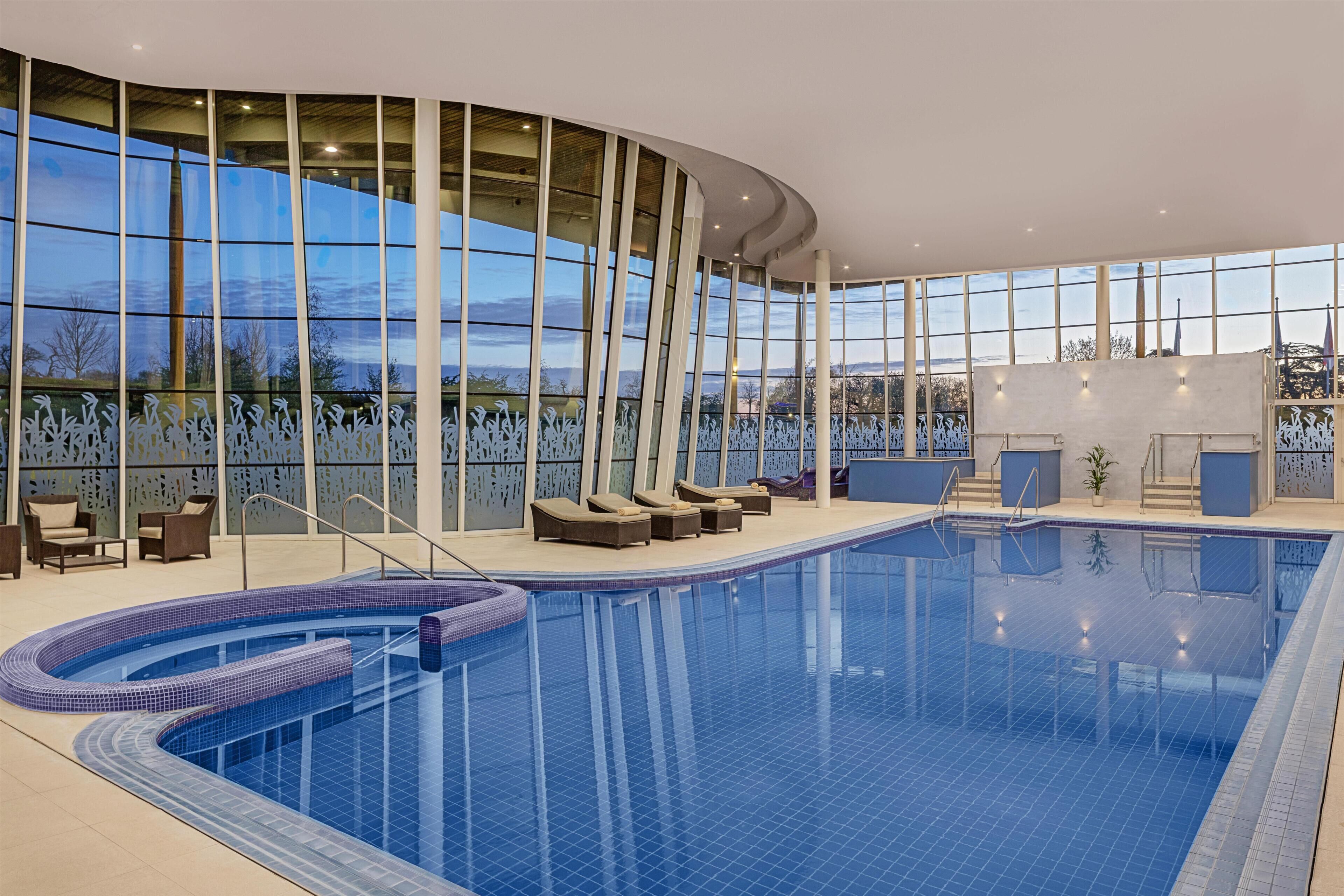 Une grande piscine intérieure à bords incurvés, entourée de chaises longues et dotée d’un mur de verre offrant une vue panoramique.
