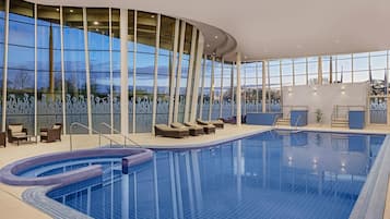 Une grande piscine intérieure à bords incurvés, entourée de chaises longues et dotée d’un mur de verre offrant une vue panoramique.