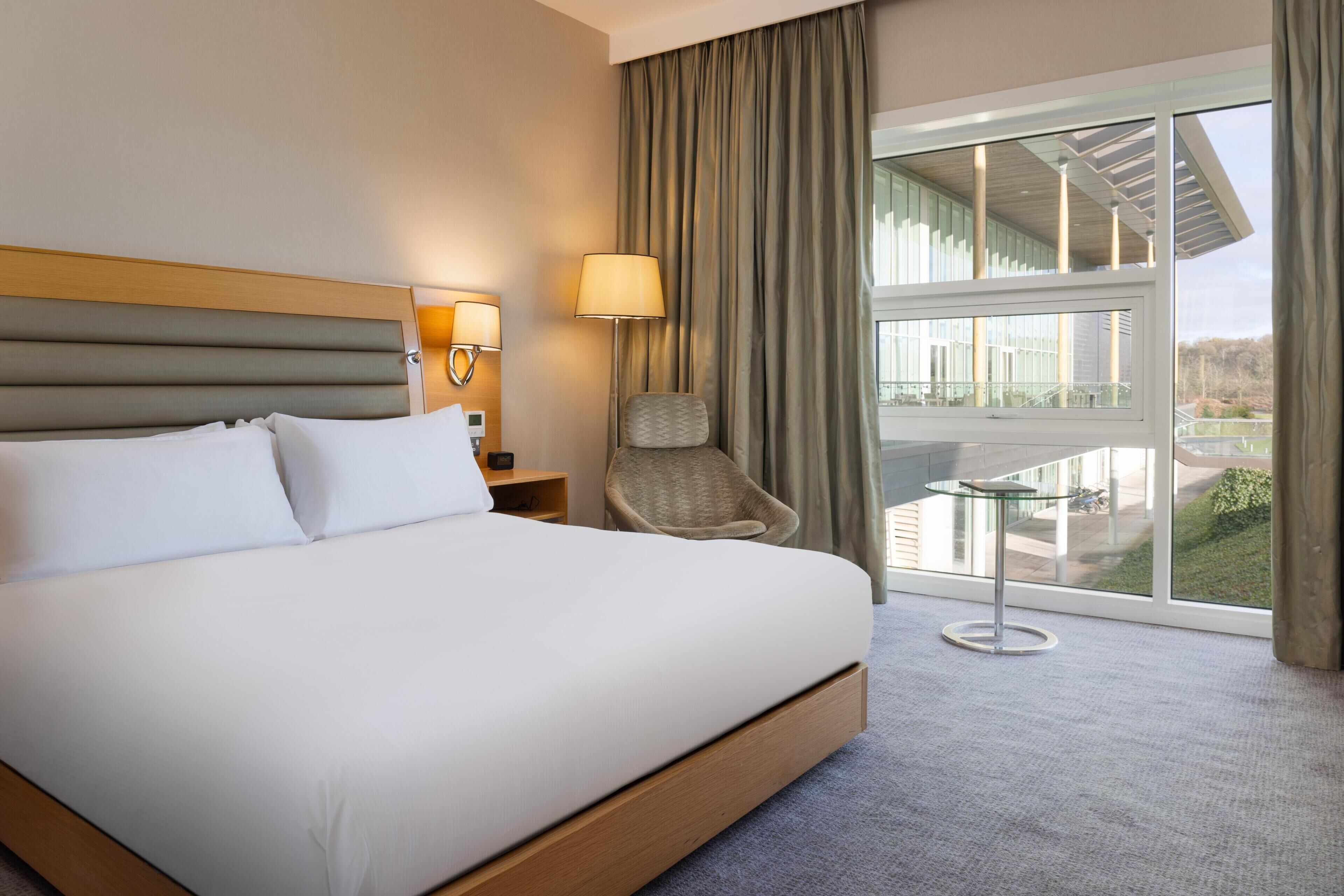 A hotel room with a large bed, a chair, a glass table, and a view of the outdoors.
