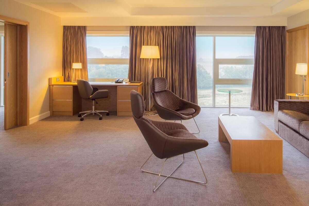 Une chambre d’hôtel équipée d’un bureau, de deux chaises, d’un canapé, d’une télévision et de grandes fenêtres.