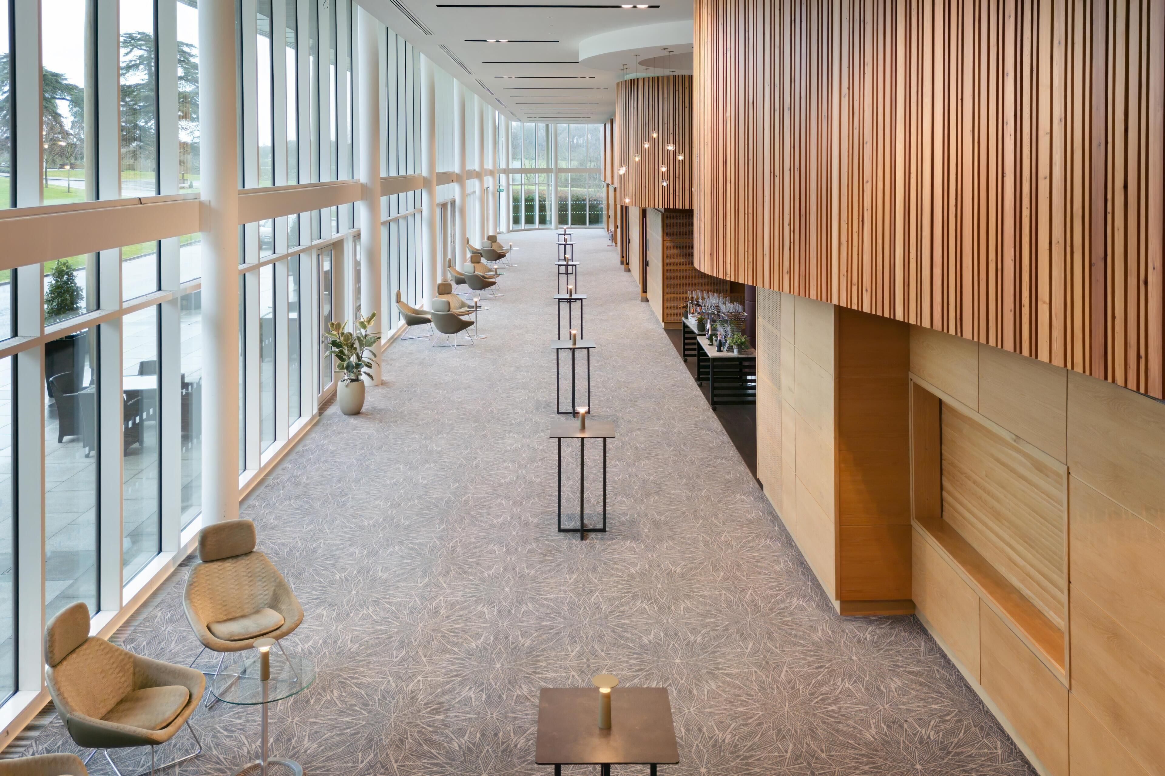 A corridor with chairs and tables, large windows, and wooden paneling.