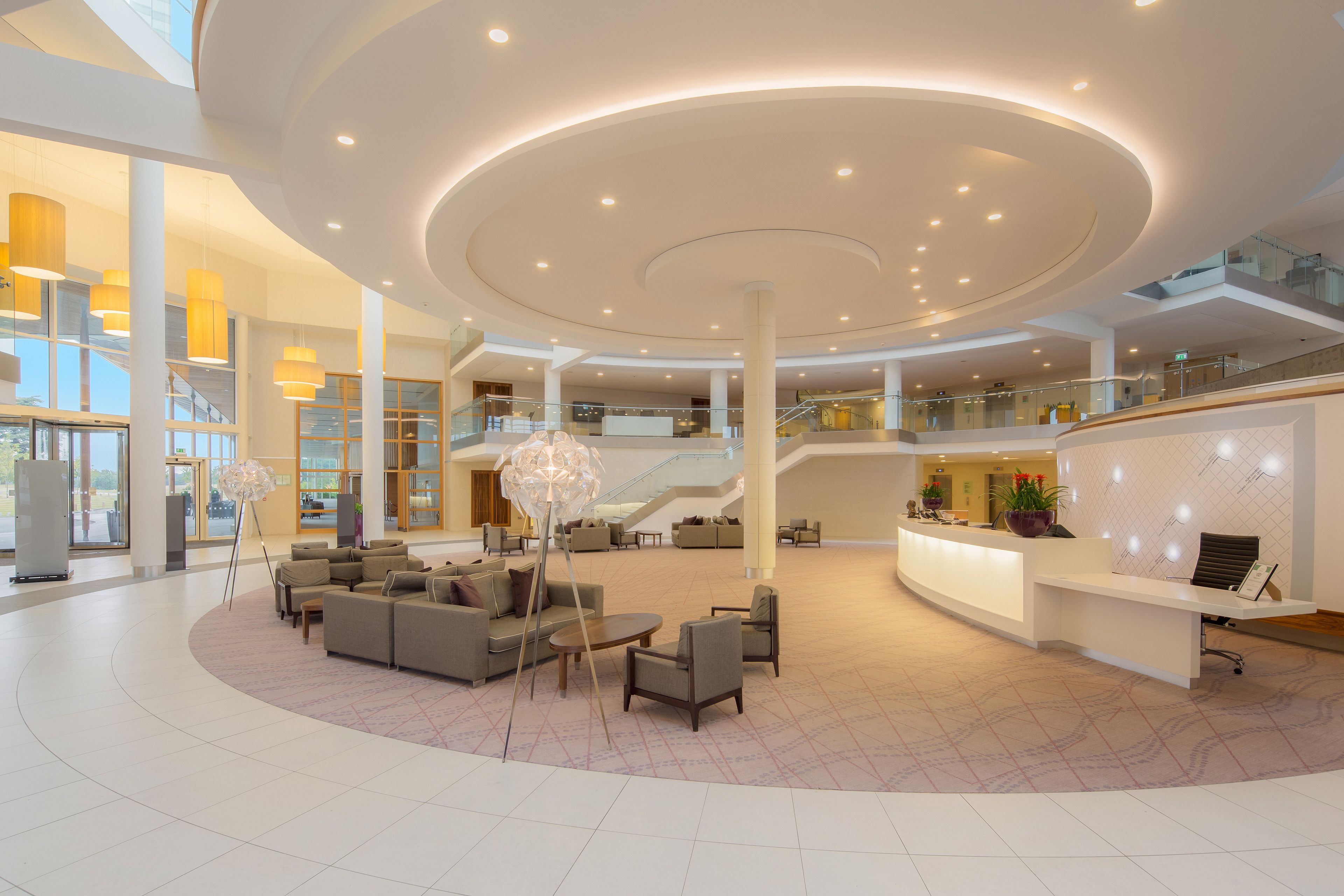Een ruime lobby met een centrale zithoek, een receptiebalie en een trap naar een hoger gelegen verdieping.