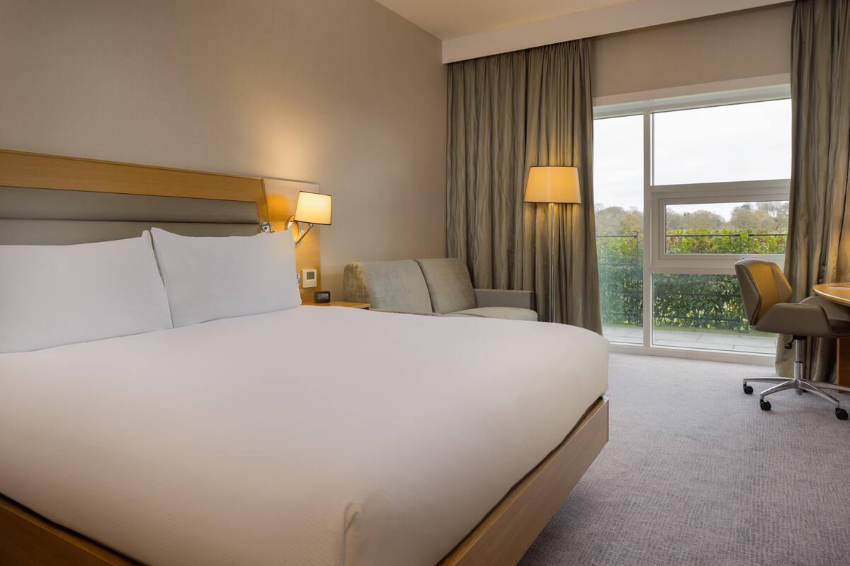 Une chambre d’hôtel avec un grand lit, un bureau, deux chaises, une fenêtre avec des rideaux et une vue sur la verdure.