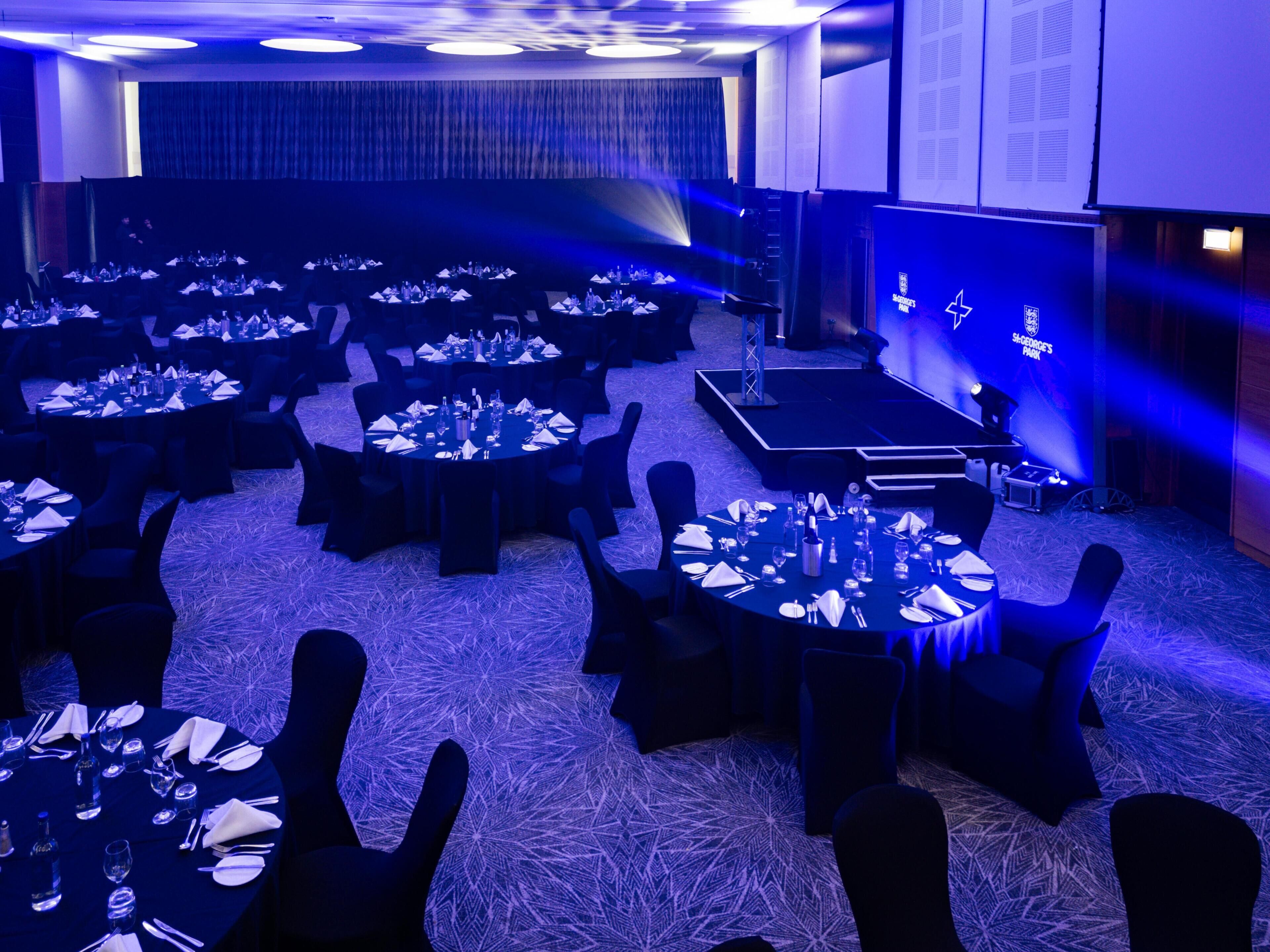 Un salón de conferencias con varias mesas dispuestas para una reunión, un escenario con un atril y una gran pantalla mostrando texto.