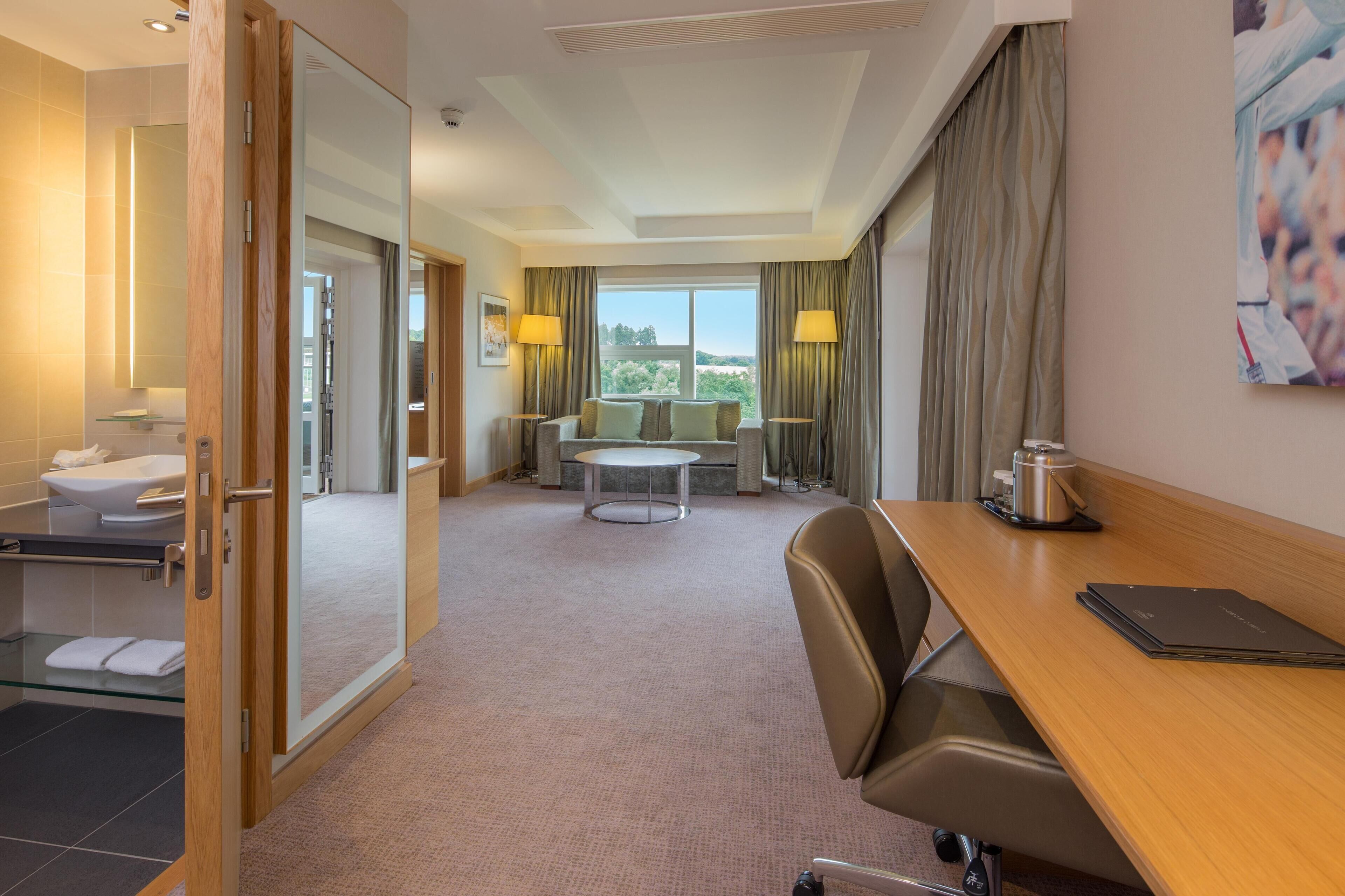 A hotel room with a wooden desk, a chair, a round table with two chairs, a glass shower enclosure, and a large window with curtains.