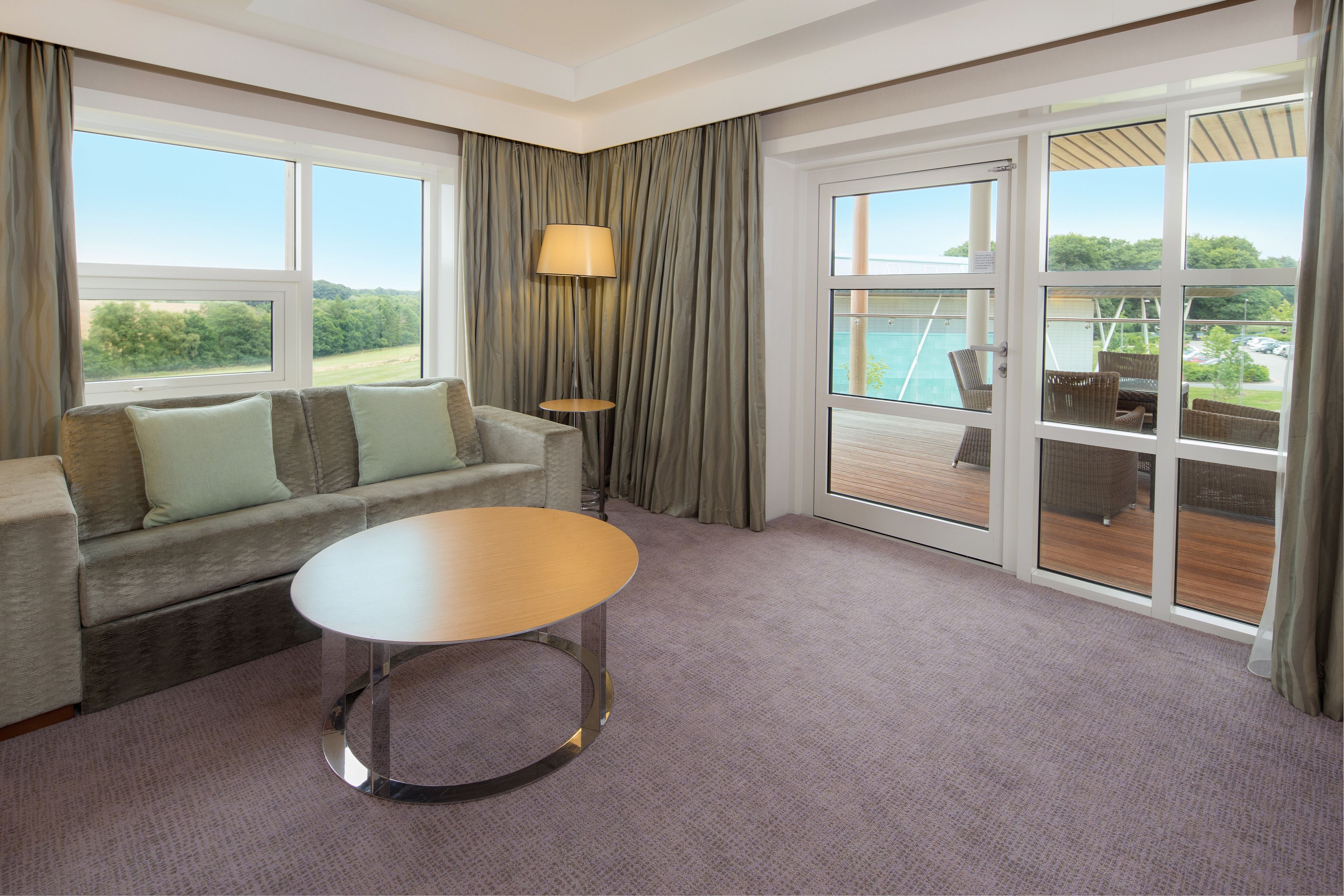 A modern hotel room with a sofa, a round table, and large windows offering a view of the outdoors.
