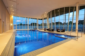 Une piscine intérieure avec des parois en verre transparent, un sol carrelé, ainsi que plusieurs chaises longues et tables.