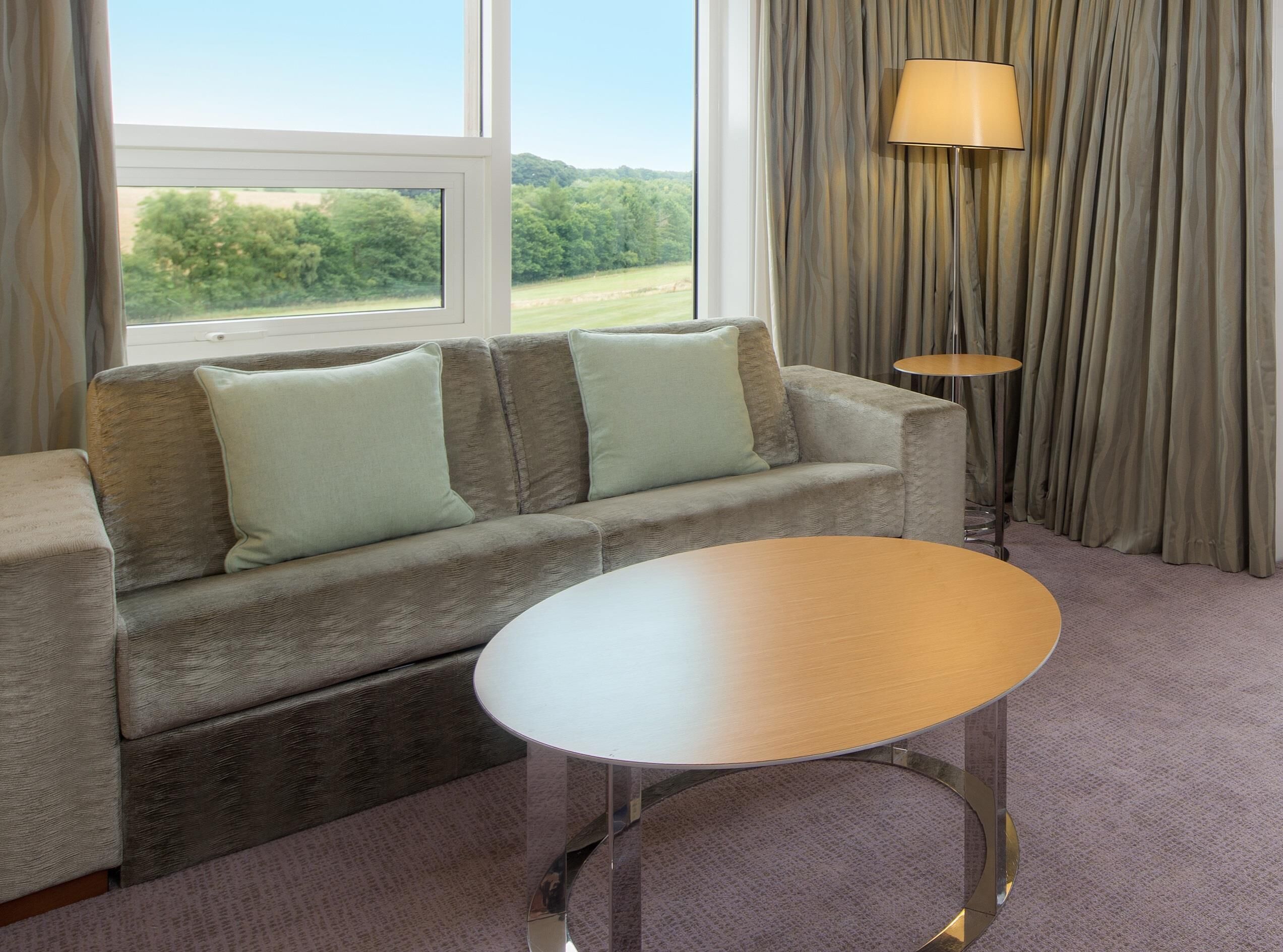 Una habitación con sofá, una mesa redonda, una lámpara y vistas al verdor a través de una ventana.