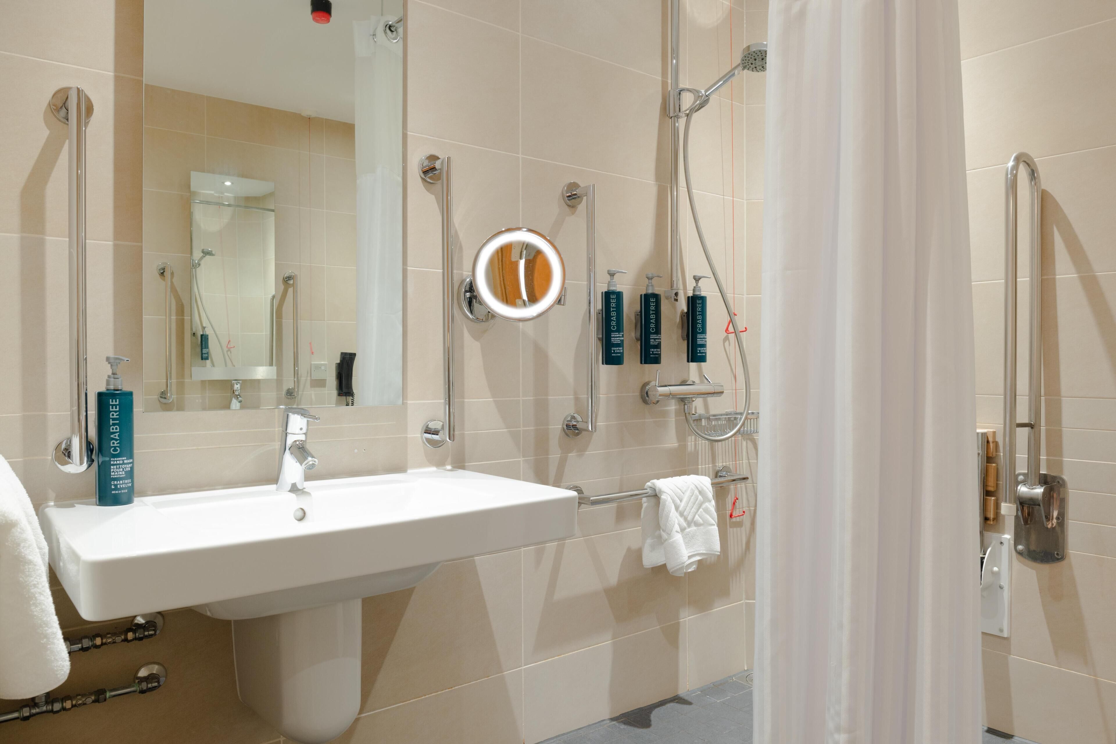 A bathroom with a sink, mirror, shower, and handrails.