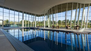A large indoor swimming pool with clear glass walls, surrounded by seating areas and a view of the outdoors. - Hilton at St George's Park, Burton Upon Trent (Burton on Trent)