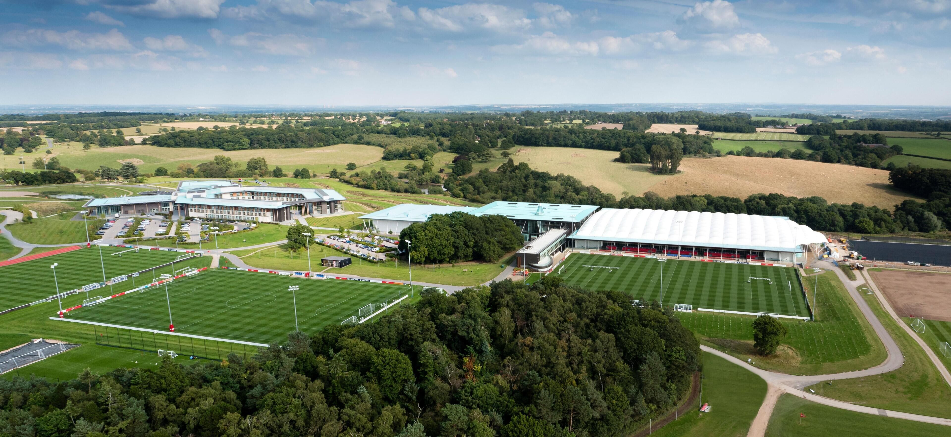 An aerial view of a sports complex with multiple fields, a large building with a green roof, and surrounding green fields.