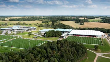 An aerial view of a sports complex with multiple fields, a large building with a green roof, and surrounding green fields.