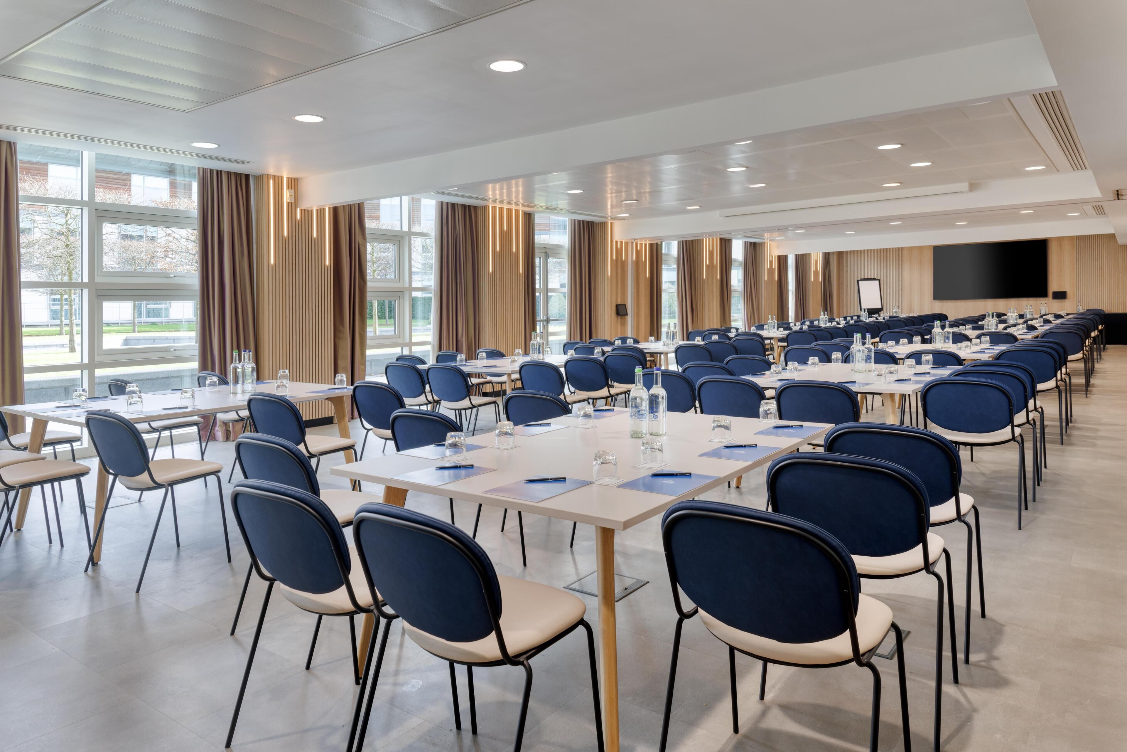 Un salón de conferencias con varias mesas y sillas, ventanas amplias y una pantalla de proyección.