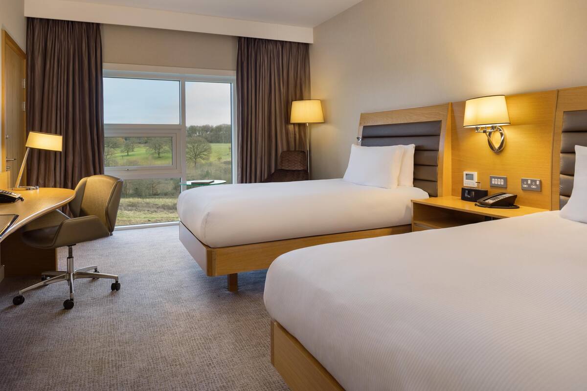 Une chambre d’hôtel avec deux lits, un bureau, une chaise, une lampe, un téléphone et une fenêtre avec des rideaux.
