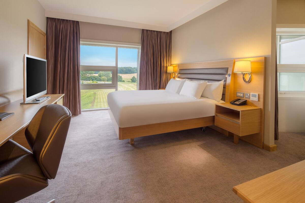 Une chambre d’hôtel comprenant un lit, un bureau avec un ordinateur, une chaise, une table de chevet avec une lampe et une fenêtre donnant sur la campagne.