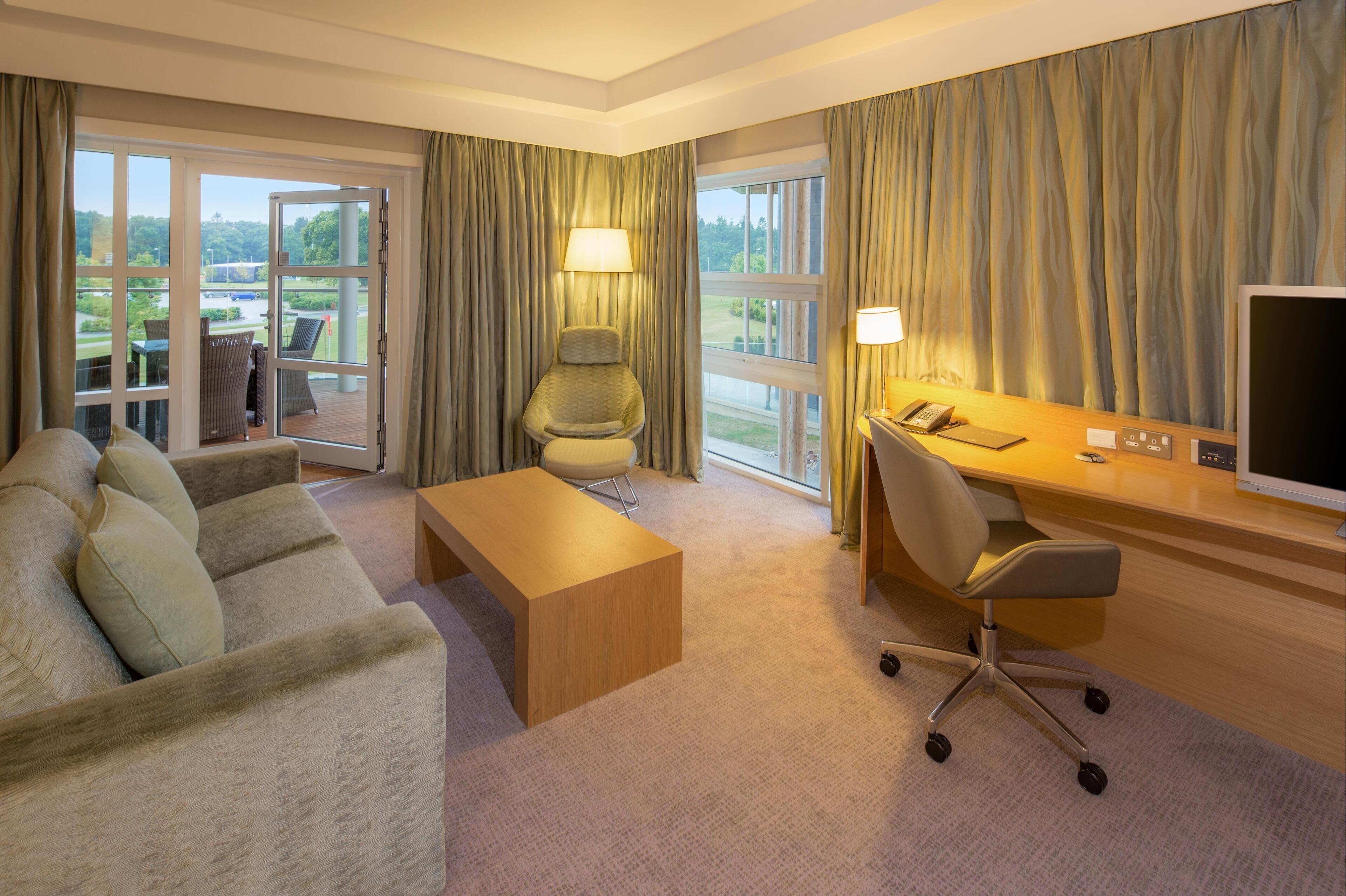 A hotel room with a sofa, a desk, a chair, a television, and a view of the outdoors.